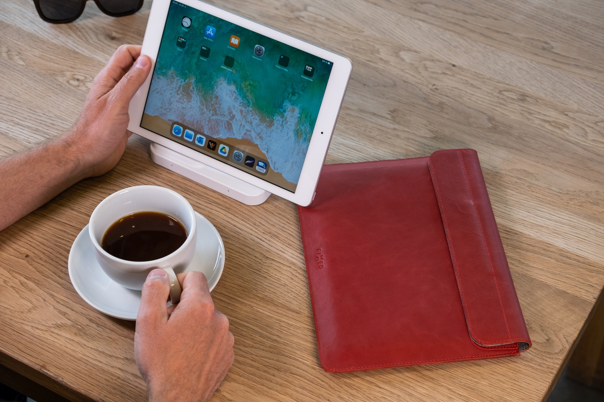Ein Mann hält eine Kaffeetasse, während sein Tablet auf einem Ständer und eine rote Hülle auf dem Holztisch liegen.