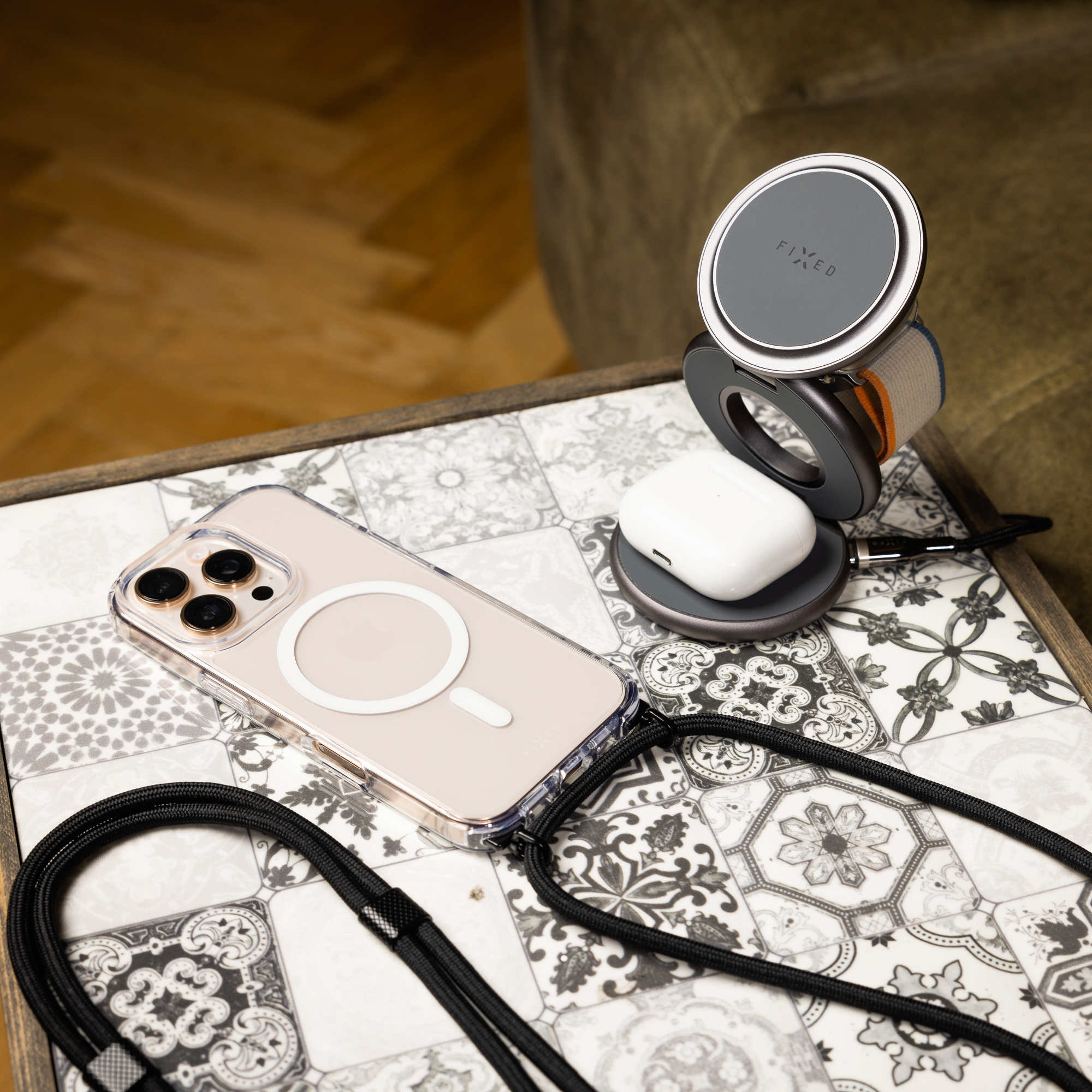Ein Smartphone in Schutzhülle mit Band und ein drahtloses Ladegerät mit Kopfhörern auf einem gefliesten Tisch.
