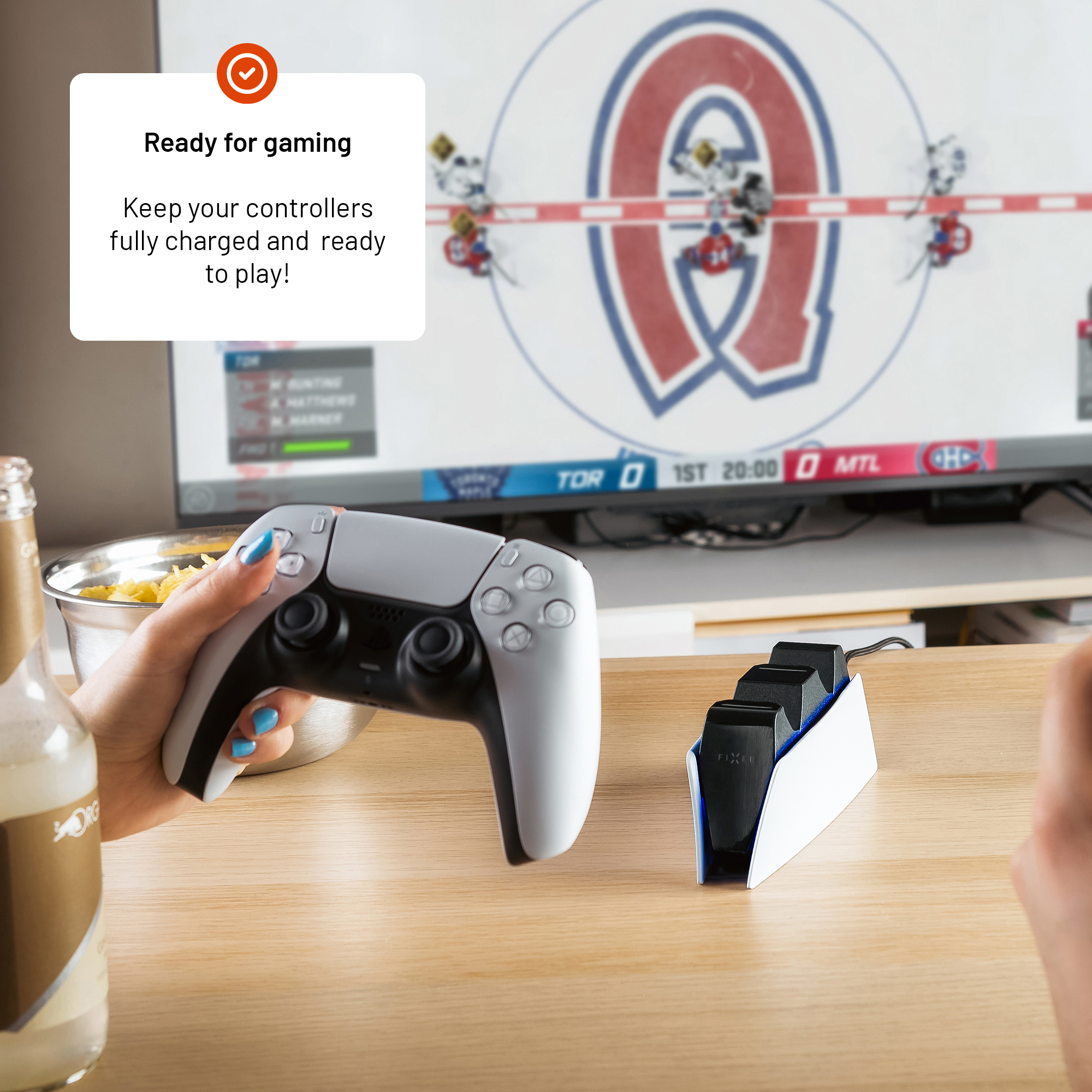 Eine Person hält einen weißen Controller in einem Raum mit Fernseher und Ladestation auf dem Tisch.
