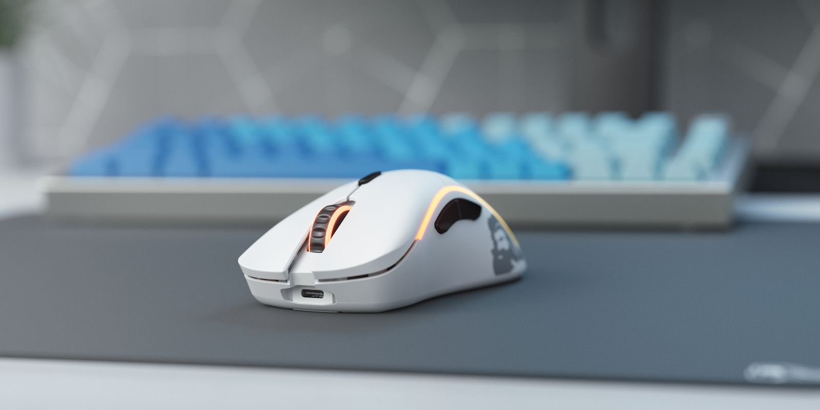 Eine weiße Gaming-Maus mit orangefarbener Beleuchtung liegt auf einem Schreibtisch vor einer blauen Tastatur.