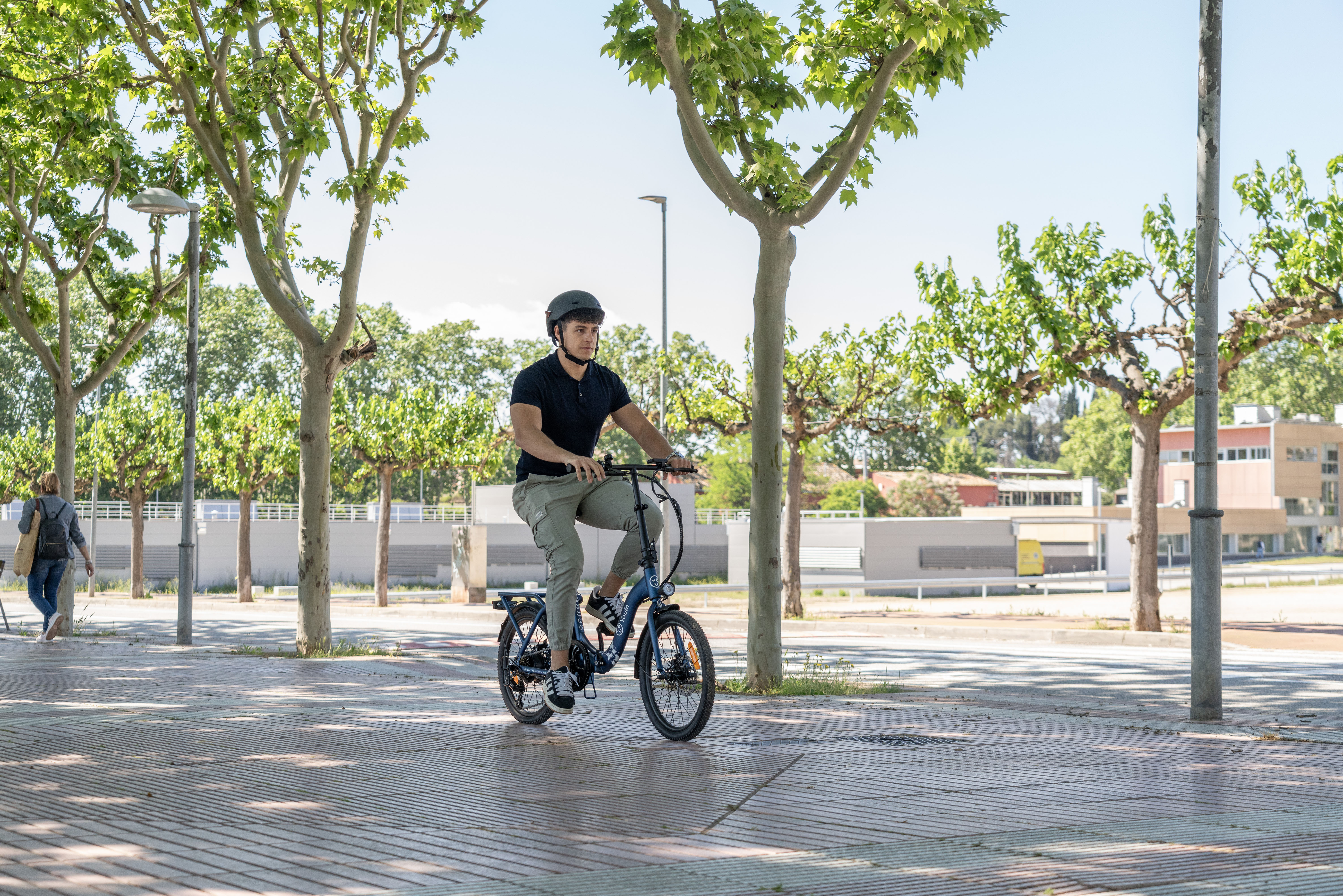 Un joven con casco monta en bicicleta eléctrica azul por un sendero soleado bordeado de árboles.