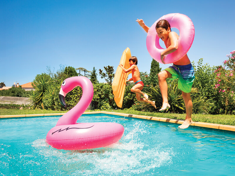 Dos niños saltan a una piscina con flotadores y una colchoneta, con un flamenco flotante en primer plano.