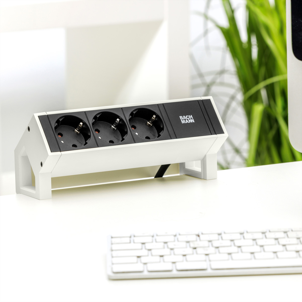 Eine weiße Steckdosenleiste mit drei schwarzen Einsätzen steht auf einem weißen Schreibtisch neben einer Tastatur.