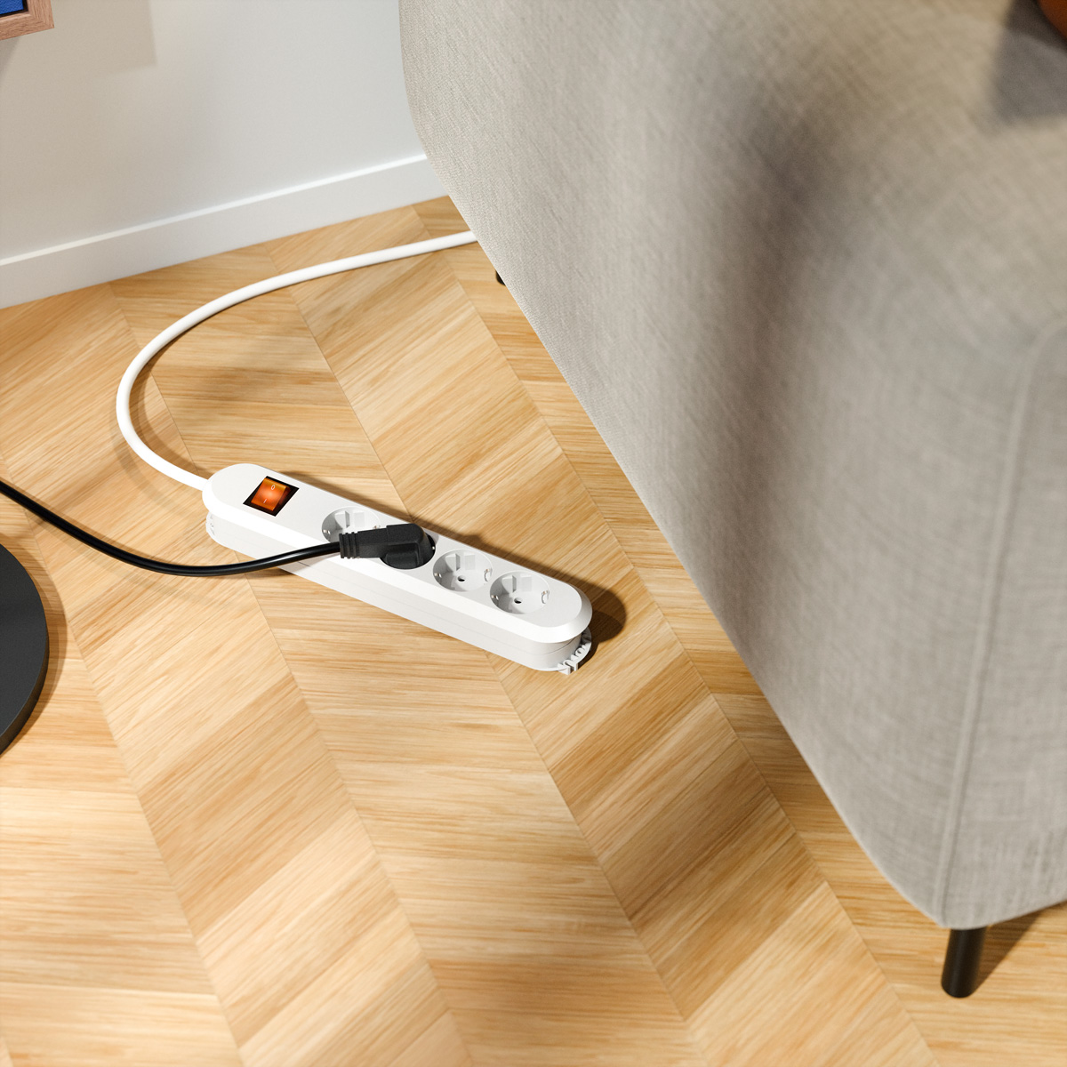 Eine weiße Steckdosenleiste mit eingestecktem schwarzem Kabel liegt auf einem Holzparkettboden neben einem grauen Sofa.