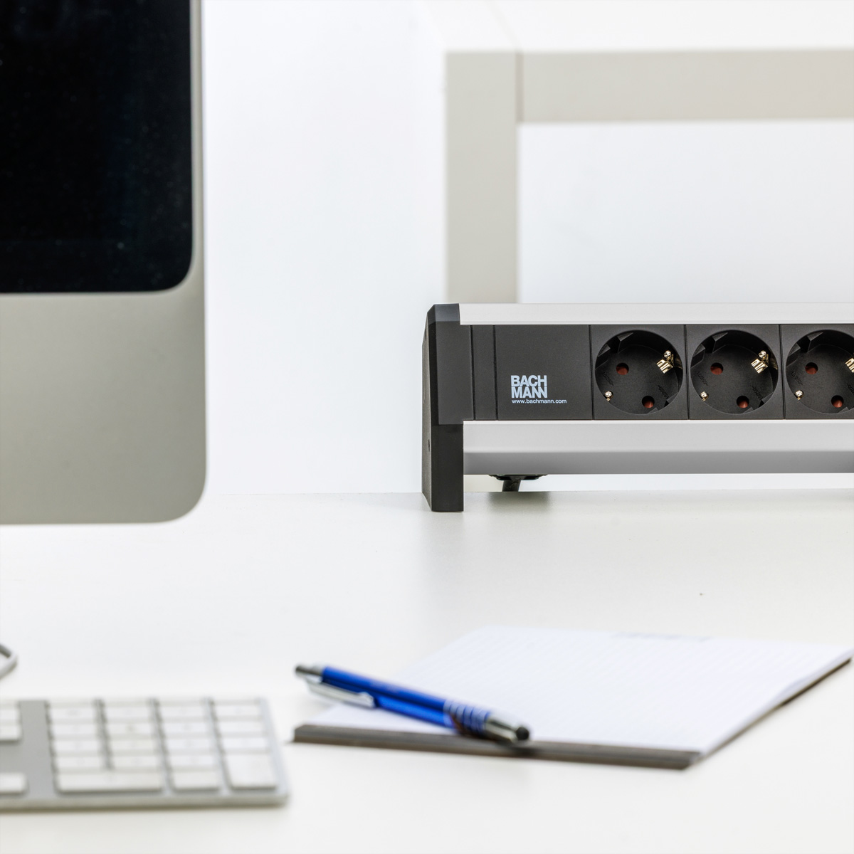 Eine Bachmann-Steckdosenleiste auf einem weißen Schreibtisch mit Computer, Tastatur, Notizblock und blauem Stift.