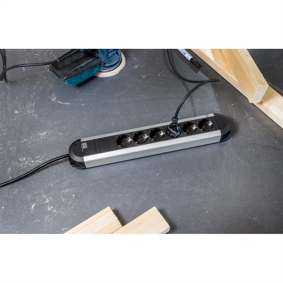 Eine Steckerleiste auf grauem Werkstattboden, umgeben von Holzstücken und einem Elektrowerkzeug.