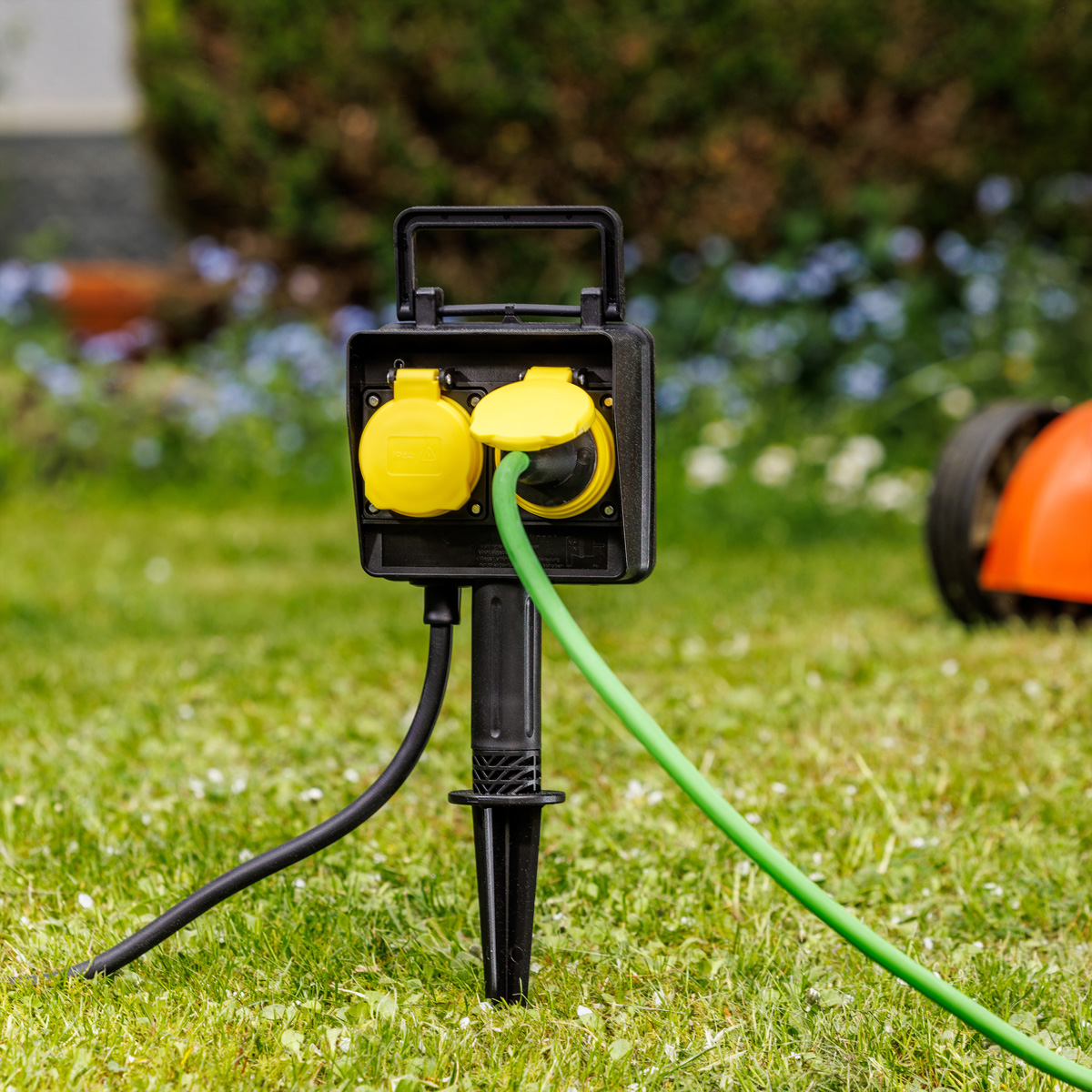 Eine schwarze Gartensteckdose mit gelben Klappdeckeln steckt im Rasen, ein grünes Kabel ist eingesteckt.