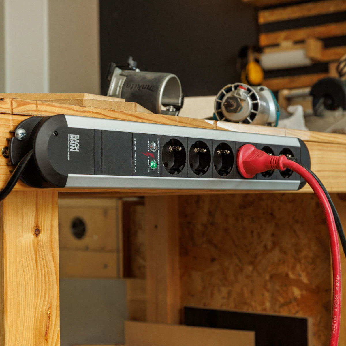 Eine schwarze und silberne Steckdosenleiste ist an einer Holzwerkbank montiert, ein rotes Kabel ist eingesteckt.