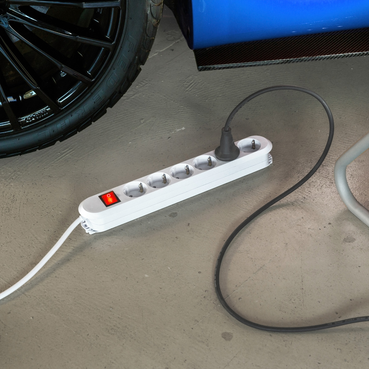 Eine weiße Mehrfachsteckdose mit eingeschaltetem rotem Schalter liegt auf dem grauen Betonboden neben einem Autoreifen.