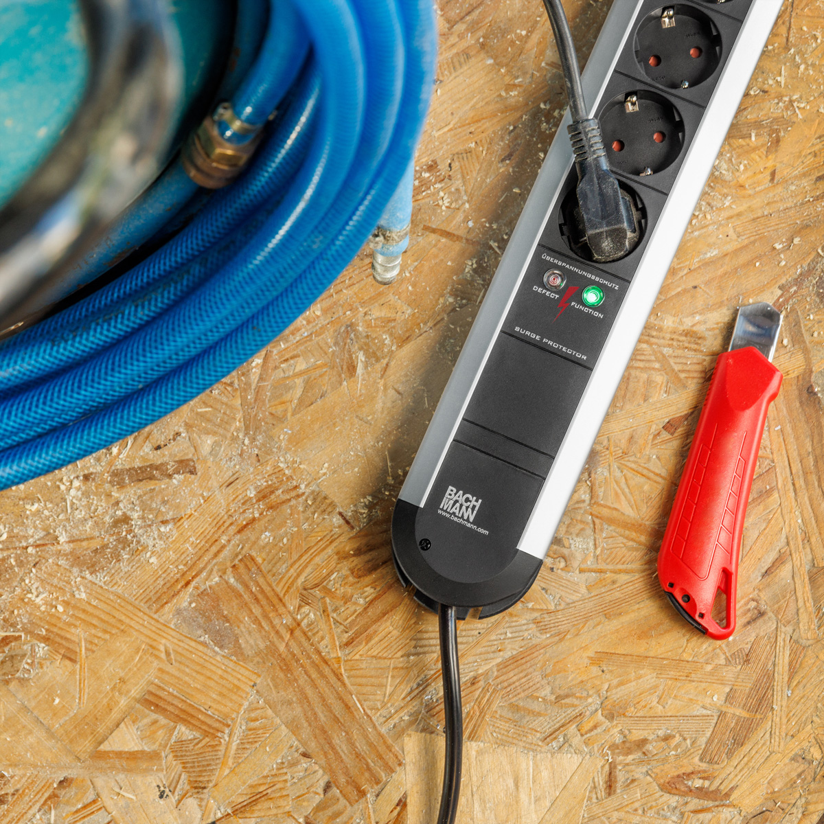 Eine Steckerleiste, ein blaues Kabel und ein rotes Teppichmesser liegen auf einer hölzernen Werkbank.
