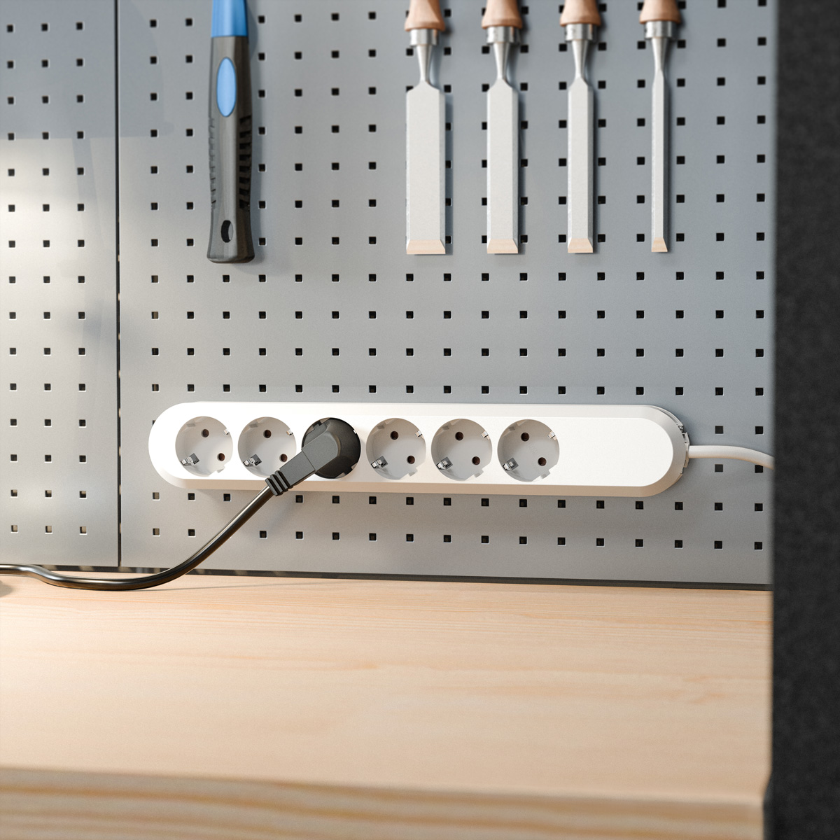 Eine weiße Mehrfachsteckdose mit eingestecktem schwarzem Kabel an einer grauen Lochwand über einem Holztisch mit Werkzeugen.