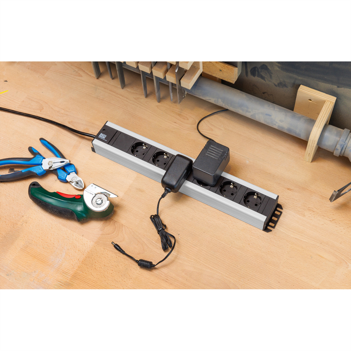 Eine Steckdosenleiste mit zwei Netzteilen liegt auf einer Holzwerkbank, daneben Pliers und ein Teppichmesser.
