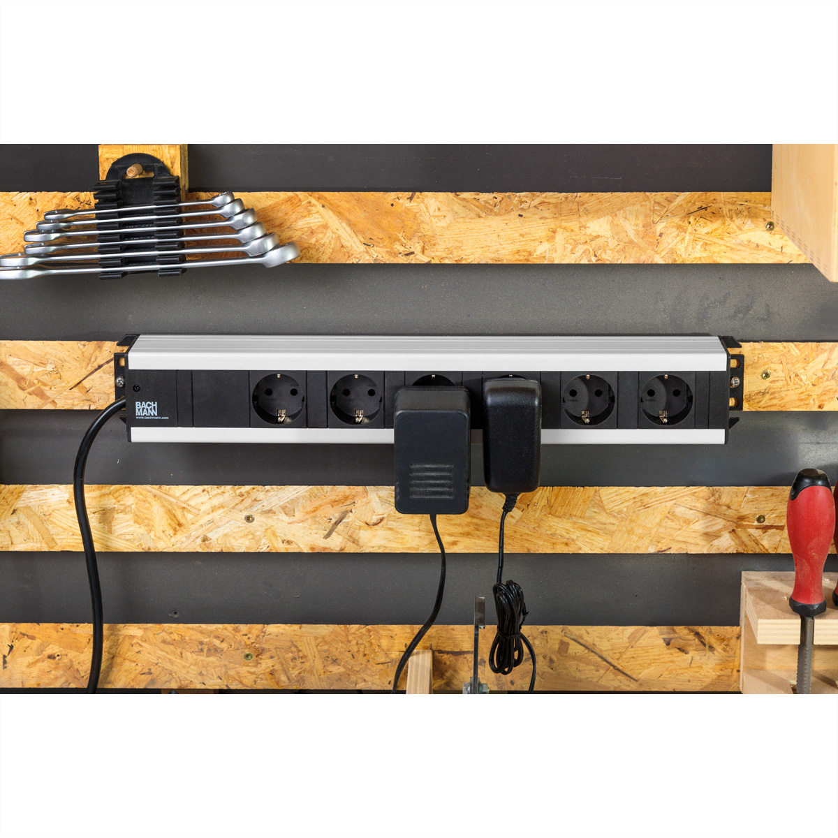 Eine schwarze und silberne Steckdosenleiste an einer Werkstattwand aus Holz und Metall, mit zwei eingesteckten Netzteilen.