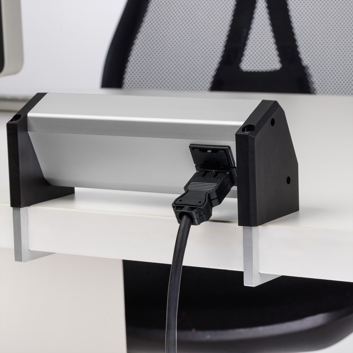 Eine graue Tischsteckdose mit schwarzen Halterungen ist an einem weißen Schreibtisch befestigt, ein schwarzes Kabel steckt.