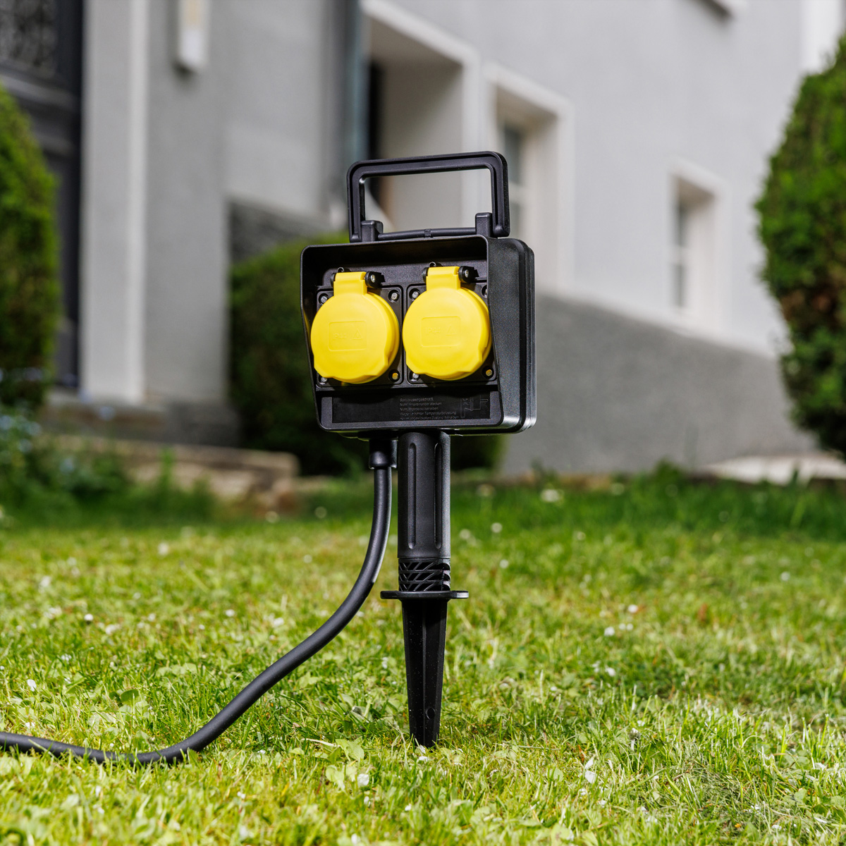 Eine schwarze Gartensteckdose mit zwei gelben Deckeln steckt im Rasen vor einem Haus.