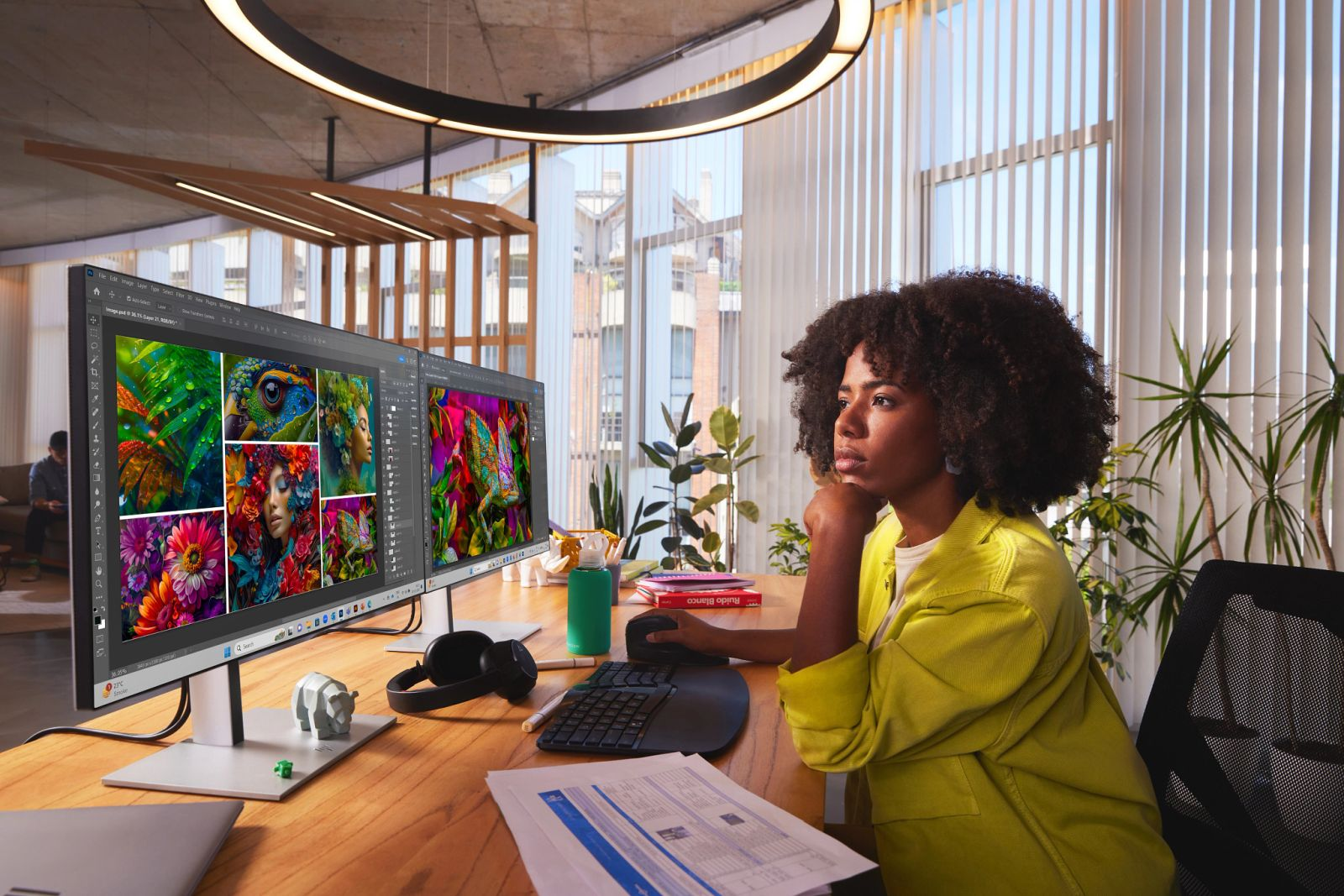 Una donna con i capelli ricci lavora a una scrivania di legno su due monitor che mostrano arte digitale.