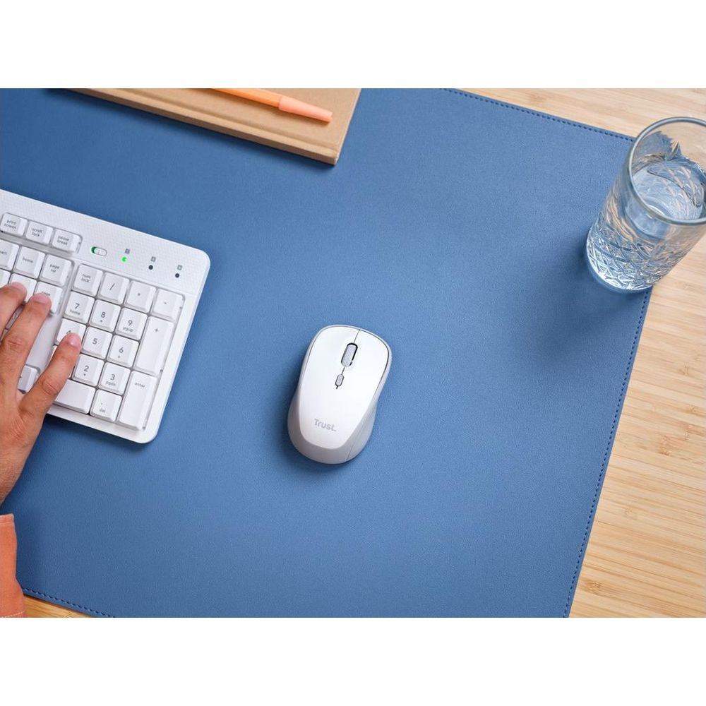 Una mano digita su una tastiera bianca accanto a un mouse Trust bianco su un sottomano blu.