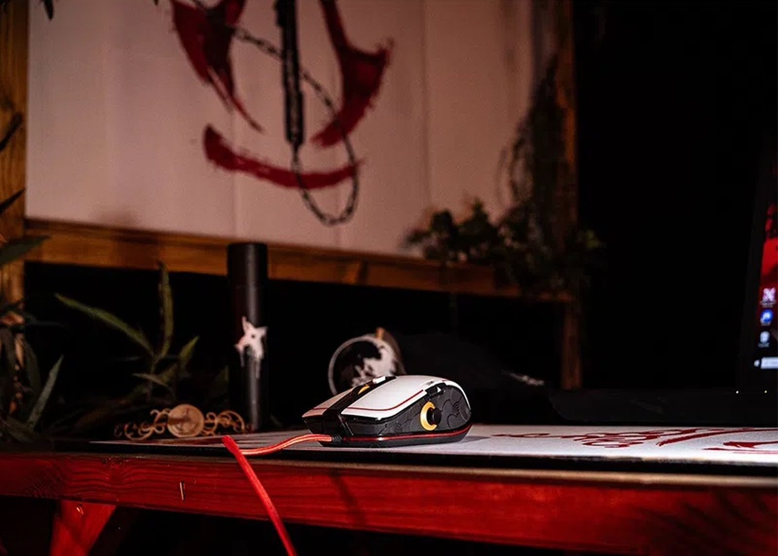 Un ratón de juego blanco y negro con cable rojo descansa en un escritorio en una habitación oscura.