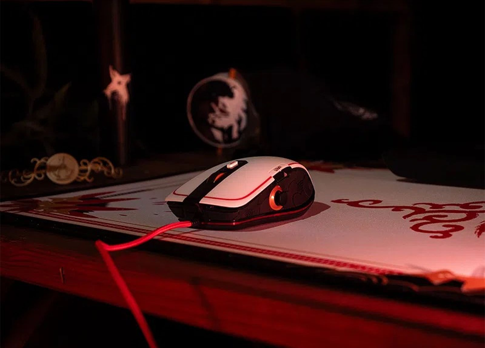 Un ratón de juego blanco y negro con cable rojo está sobre una alfombrilla roja en un entorno oscuro.
