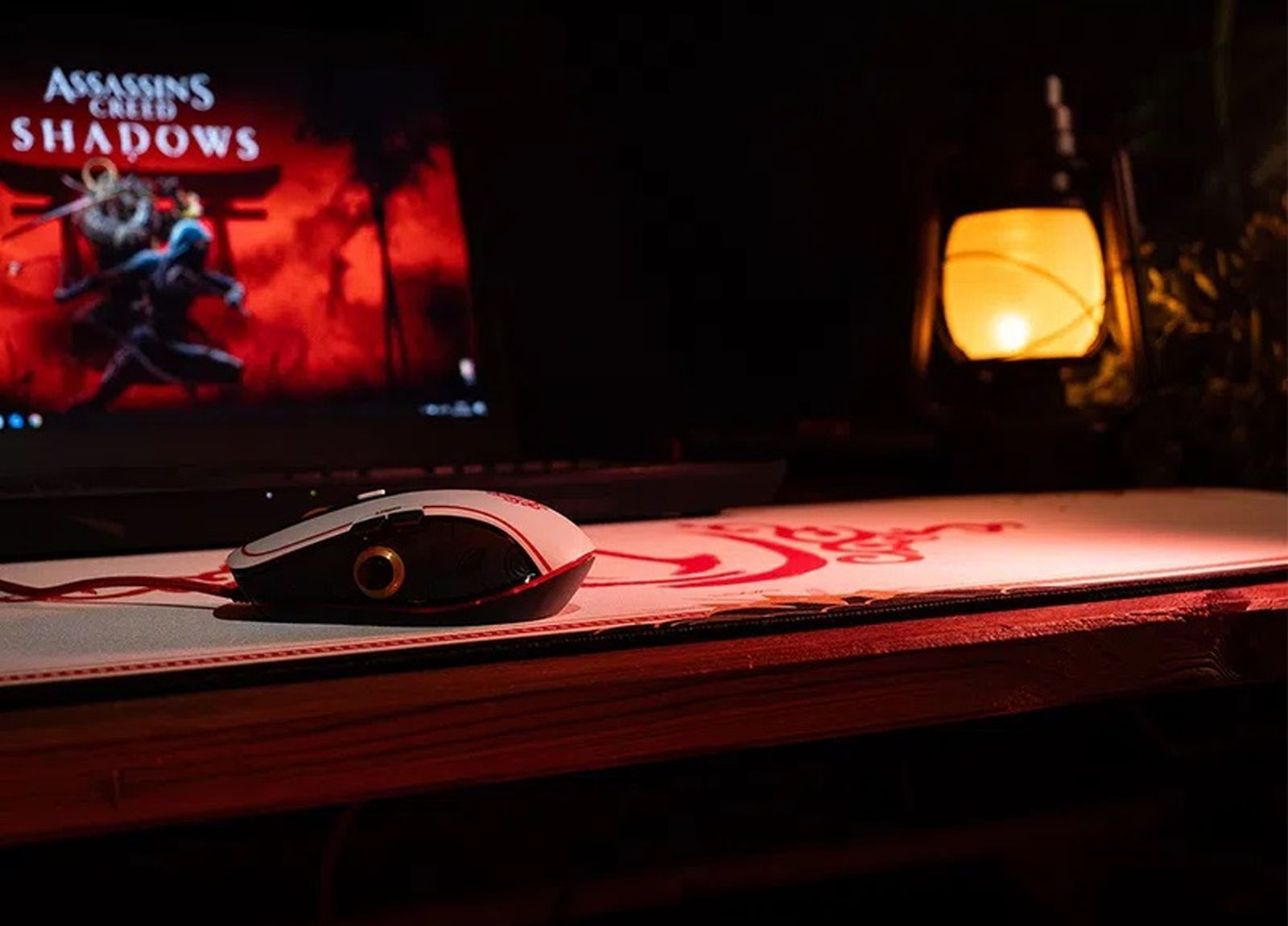 Un ratón de juego en una alfombrilla roja frente a un portátil con Assassin's Creed Shadows en una habitación oscura.