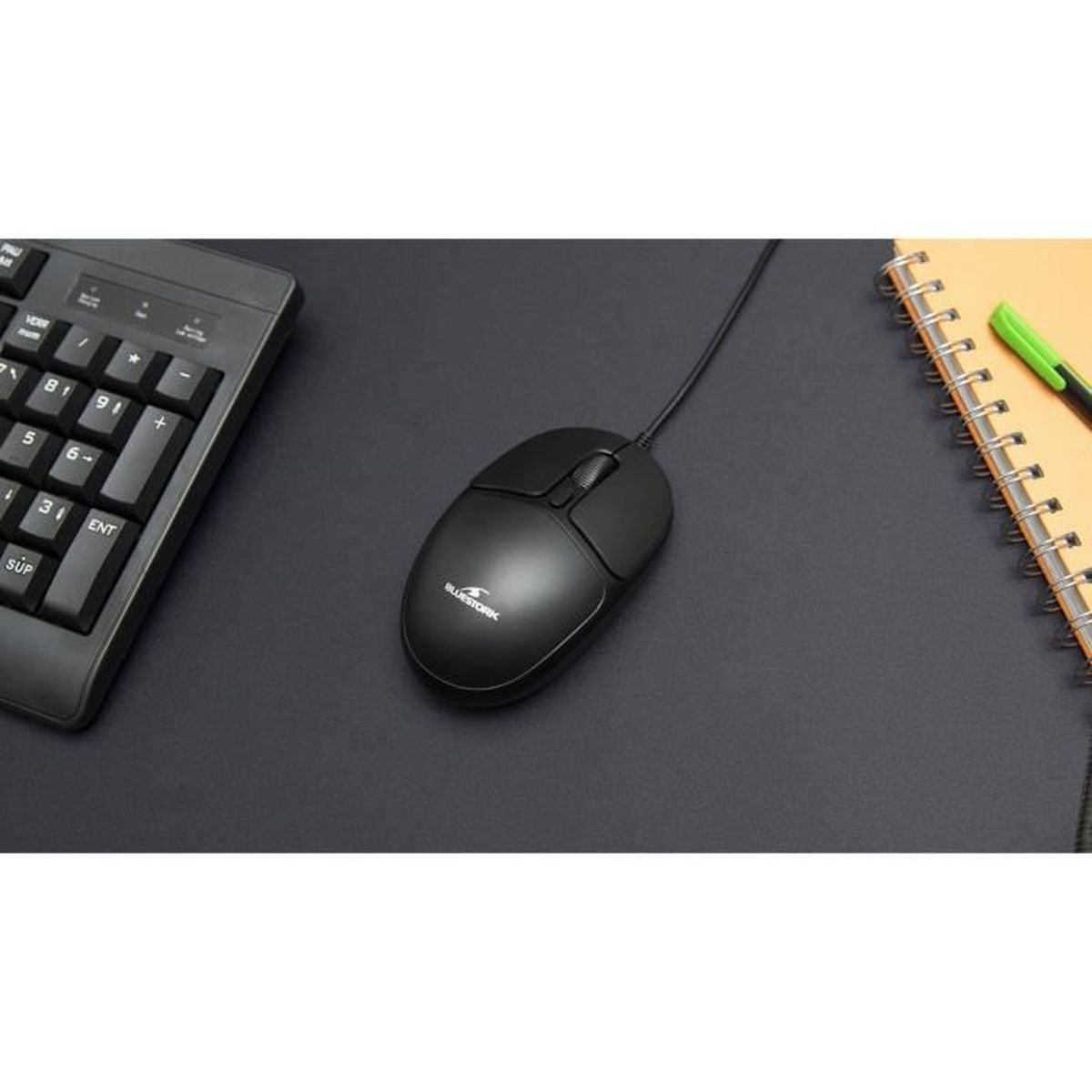Un ratón de ordenador negro en un escritorio oscuro, con un teclado a la izquierda y un bloc de notas a la derecha.