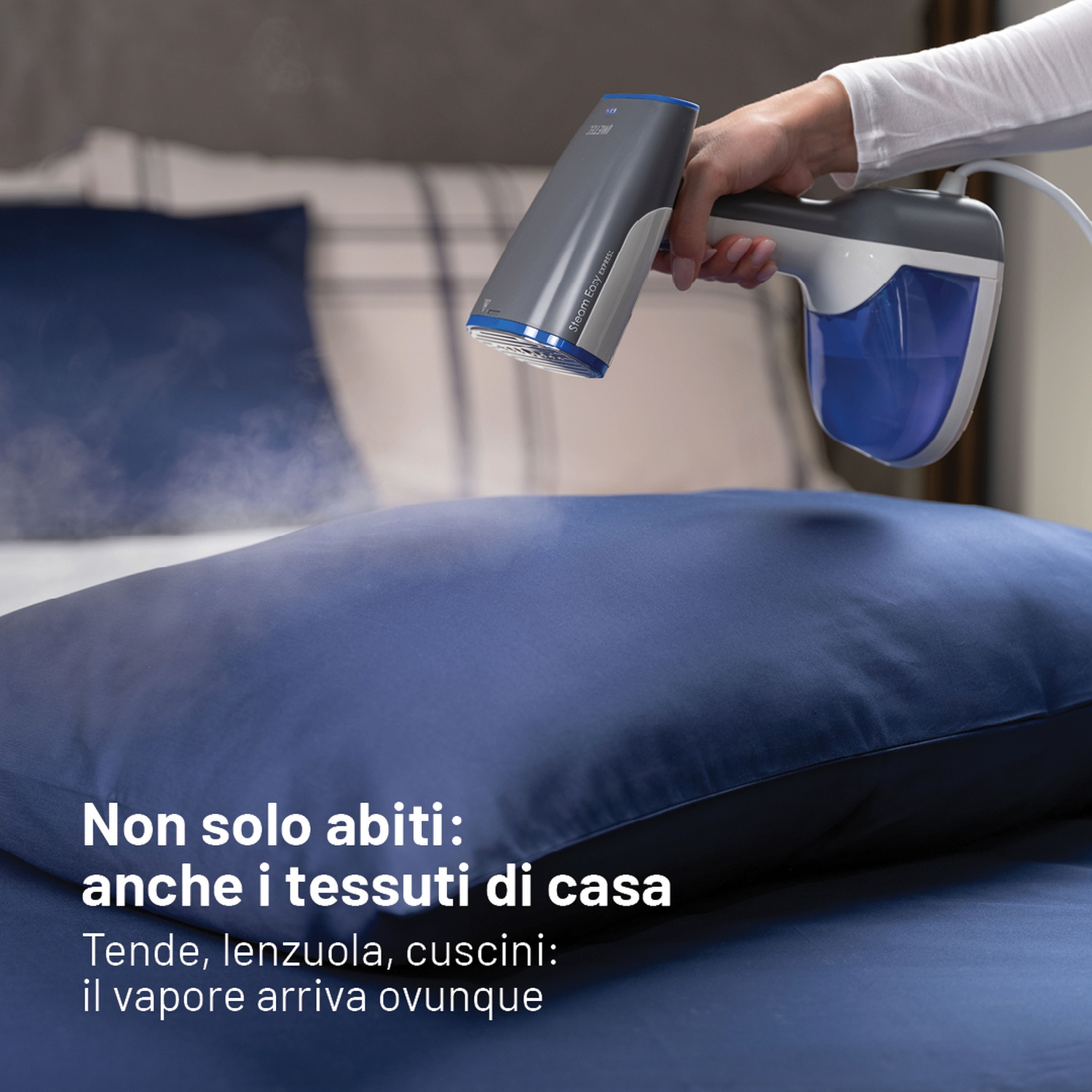 Eine Hand hält einen grauen und blauen Handdampfglätter, der Dampf auf ein blaues Kissen auf einem Bett abgibt.