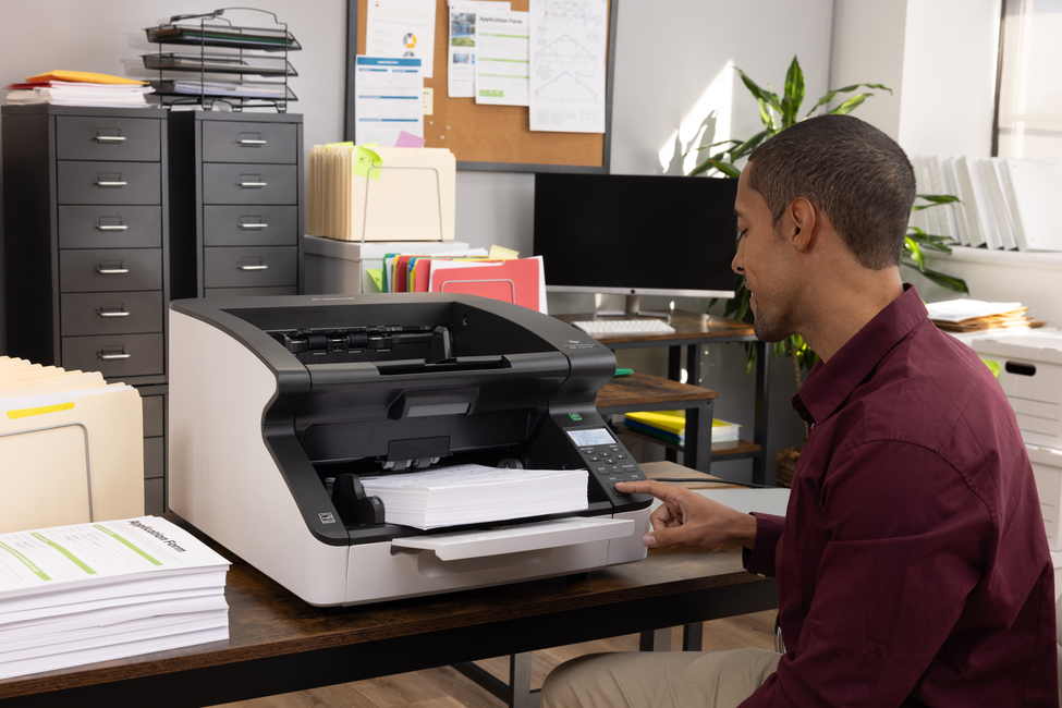 Ein Mann in einem dunkelroten Hemd bedient einen großen Dokumentenscanner in einem Büro mit Aktenschränken.