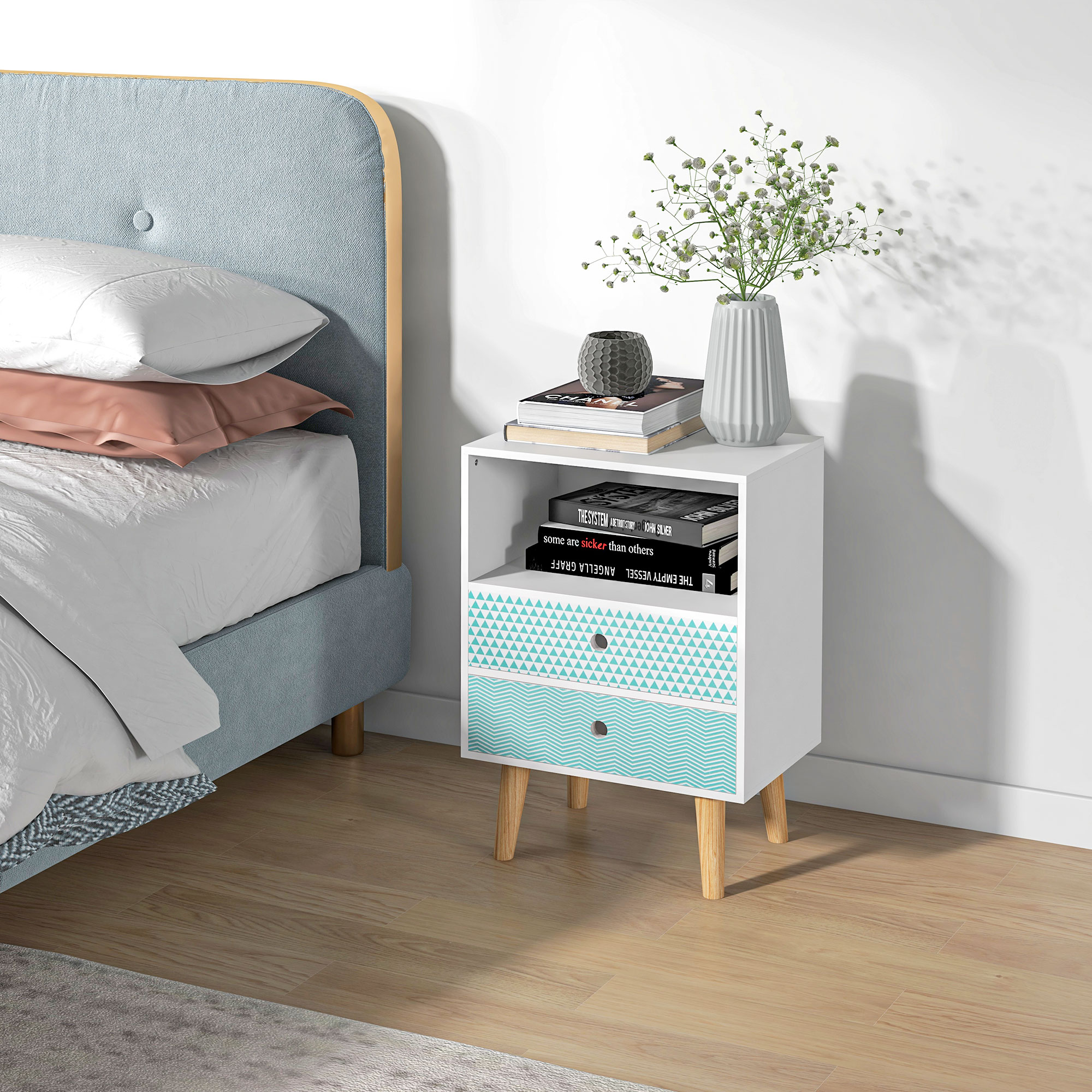 Una mesita de noche blanca con cajones estampados, libros y un jarrón con flores junto a una cama azul en suelo de madera.