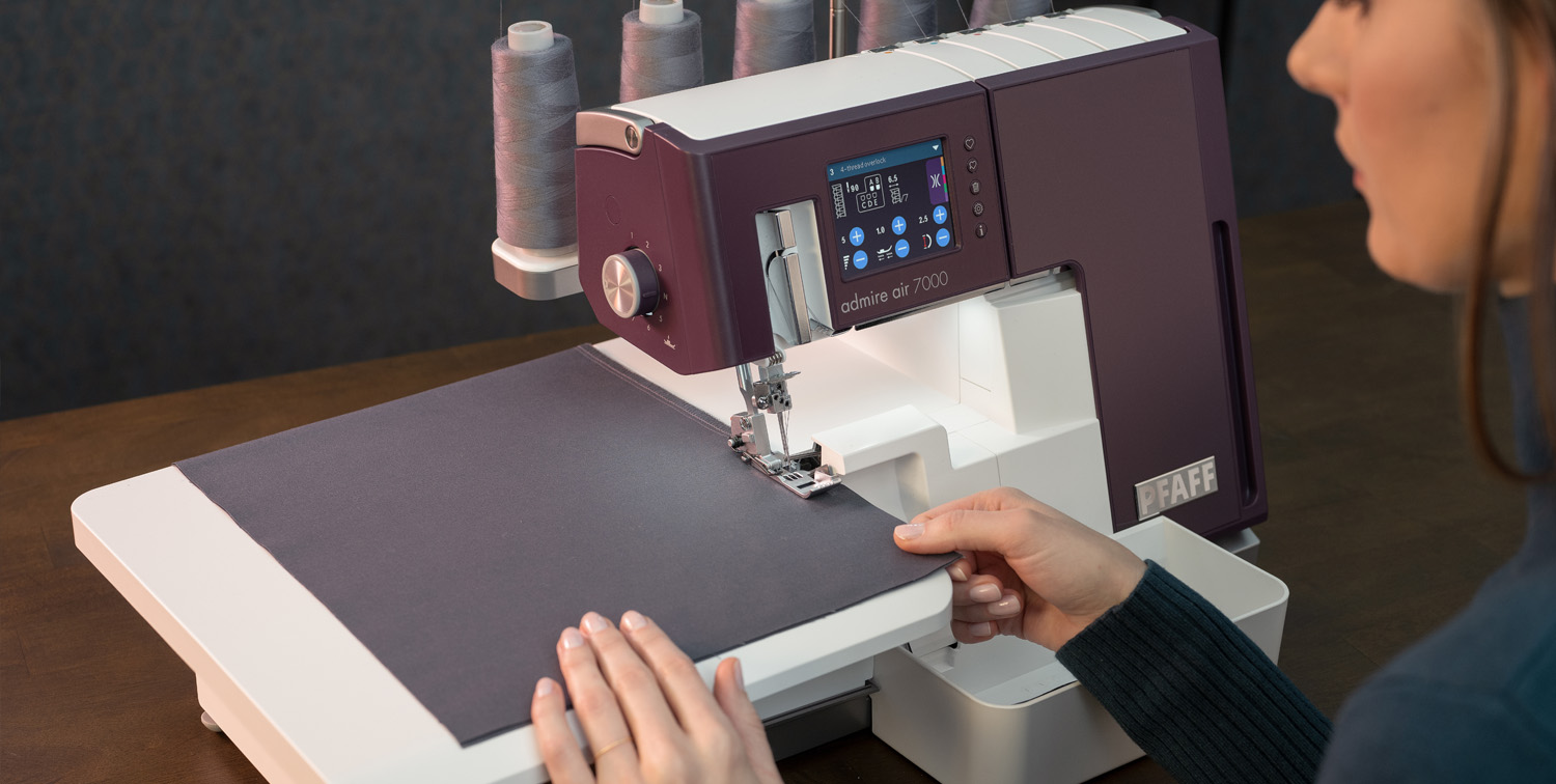 Una mujer usa una máquina de coser Pfaff blanca y morada guiando tela gris.