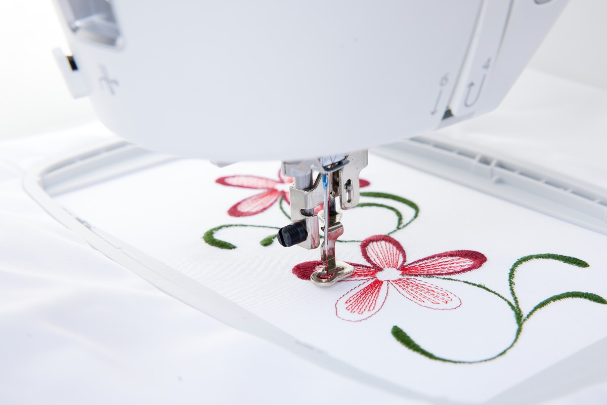 Una aguja de máquina de coser borda un patrón floral rojo y enredadera verde en tela blanca.