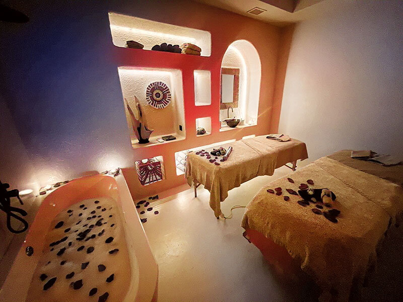 Una sala de spa con dos camillas de masaje, una bañera llena de agua y pétalos de rosa, y nichos de pared iluminados.