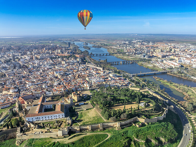 Vista aérea de una ciudad histórica con un río, un parque y un globo aerostático en el cielo azul.