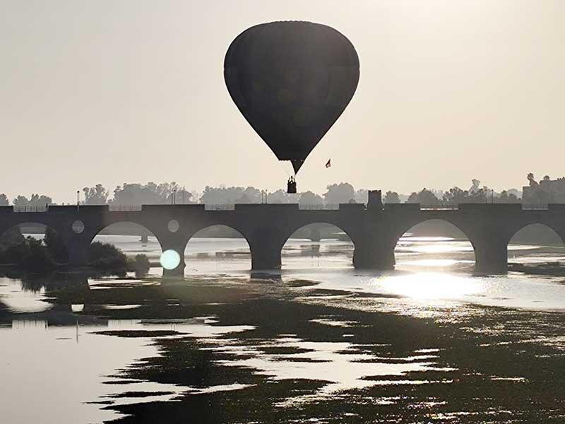 Un globo aerostático vuela sobre un puente de piedra de varios arcos sobre un río tranquilo al amanecer.