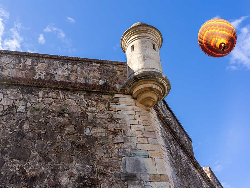 Una antigua fortaleza de piedra con torre de vigilancia bajo un cielo azul y un globo aerostático.