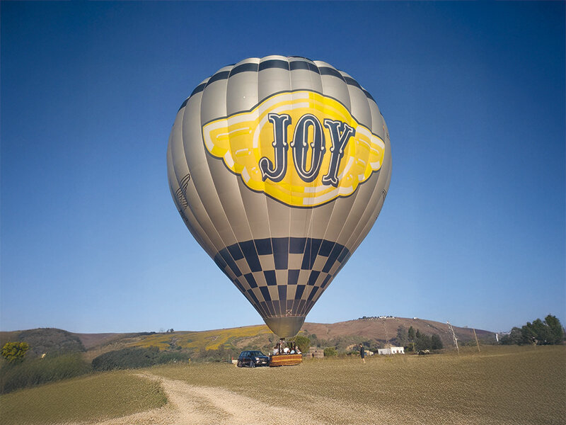 Un globo aerostático gris con el texto 'JOY' flota sobre un campo bajo un cielo azul.