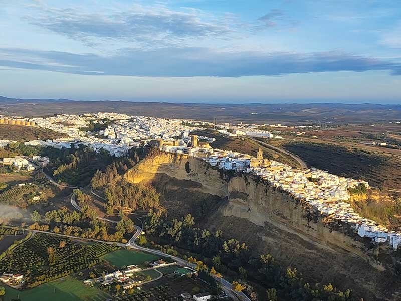 Vista aérea de un pueblo blanco sobre un acantilado, rodeado de campos y bosques bajo un cielo azul.