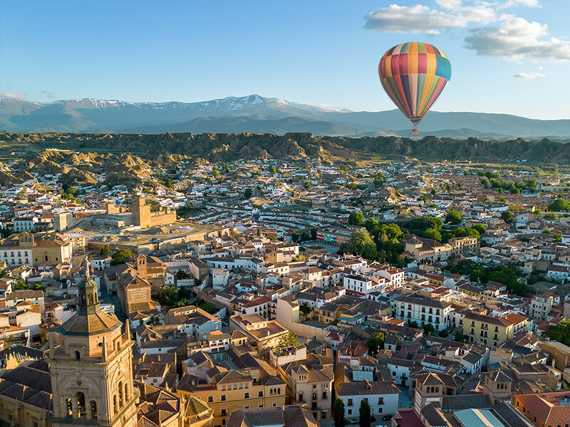 Globo aerostático colorido sobre una ciudad con montañas al fondo.