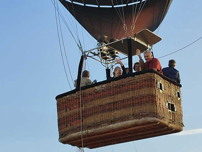 Personas saludan felices desde la cesta de un globo aerostático en el cielo.