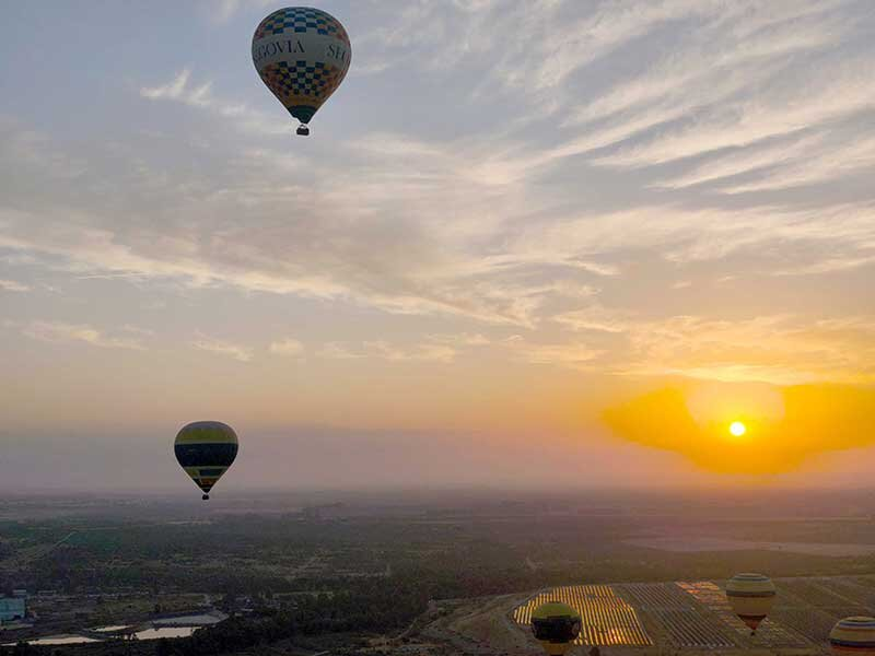 Varios globos aerostáticos vuelan sobre un amplio paisaje con campos al atardecer.