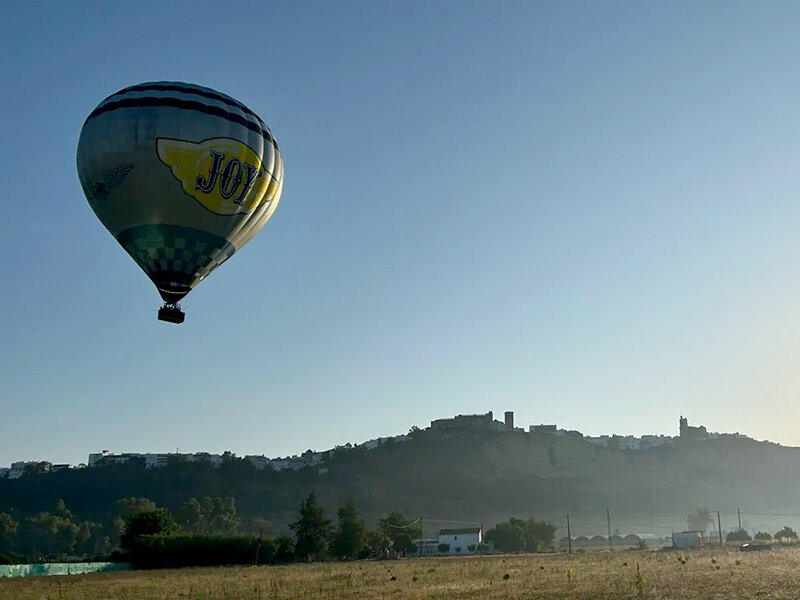 Un globo aerostático con el texto JOY vuela sobre un paisaje con edificios y campos.