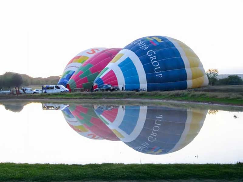 Tres coloridos globos aerostáticos desinflados yacen junto a un lago tranquilo, reflejándose en el agua.