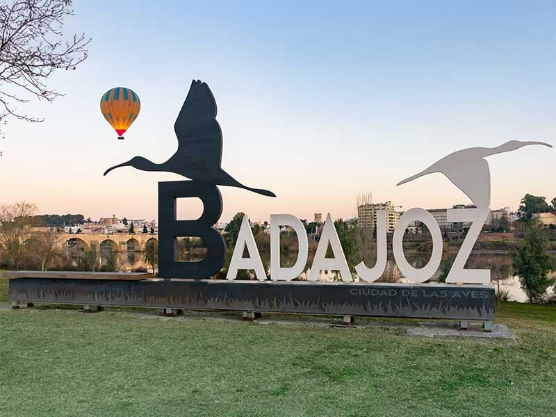 Letrero de Badajoz con cigüeñas en un parque, con globo aerostático y ciudad al fondo.