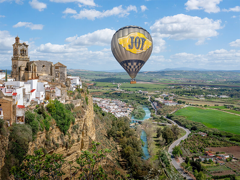 Un globo aerostático con la palabra JOY vuela sobre un pueblo histórico en un acantilado y un paisaje verde.