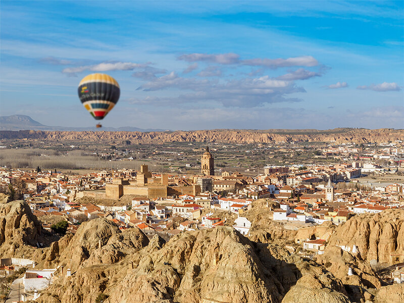 Un globo aerostático vuela sobre un pueblo con edificios históricos, rodeado de un paisaje rocoso.