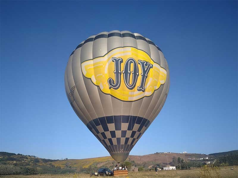Un gran globo aerostático a cuadros grises y negros con la palabra JOY en un emblema amarillo flotando sobre un paisaje.