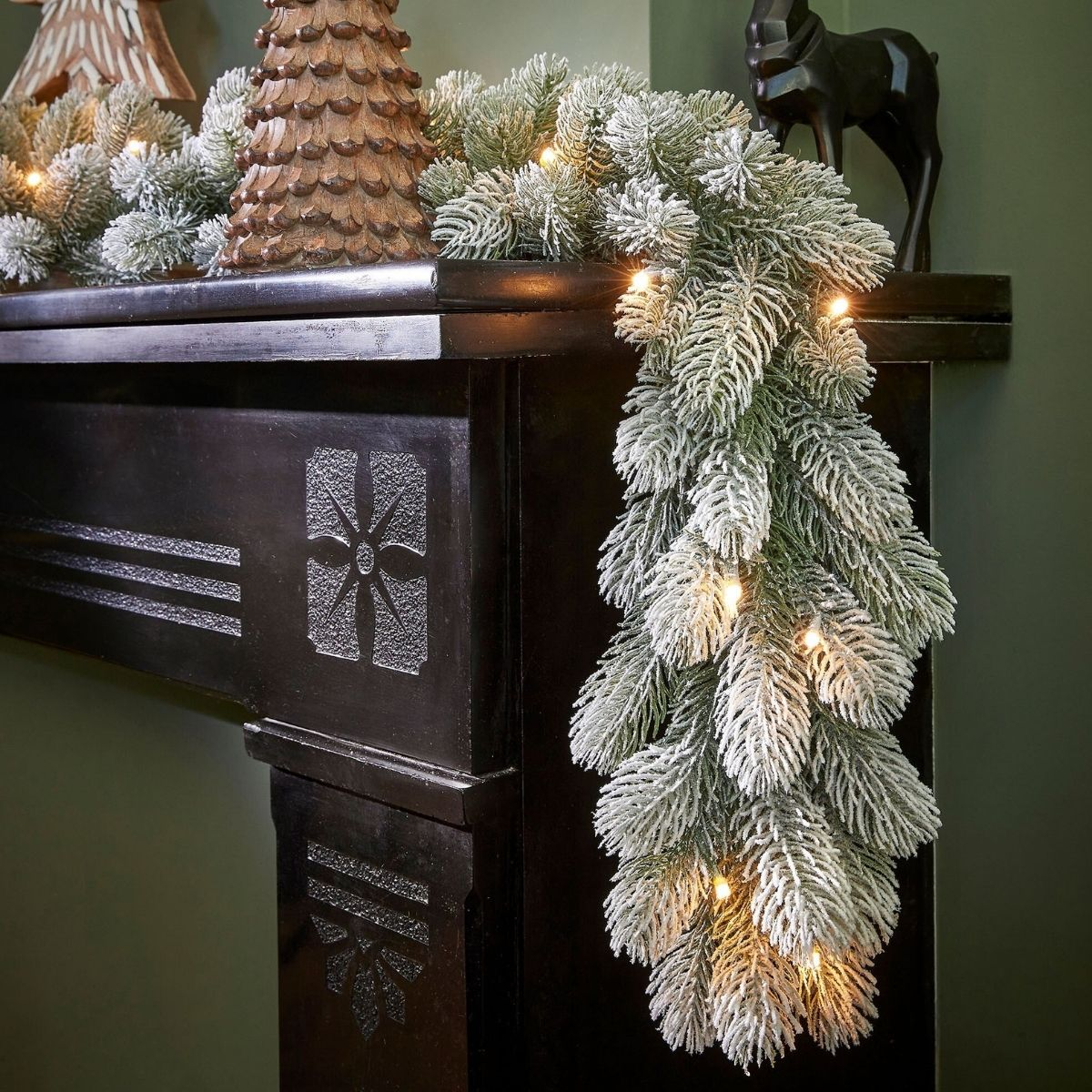 Eine verschneite Weihnachtsgirlande mit Lichtern dekoriert einen dunklen Holzkamin.