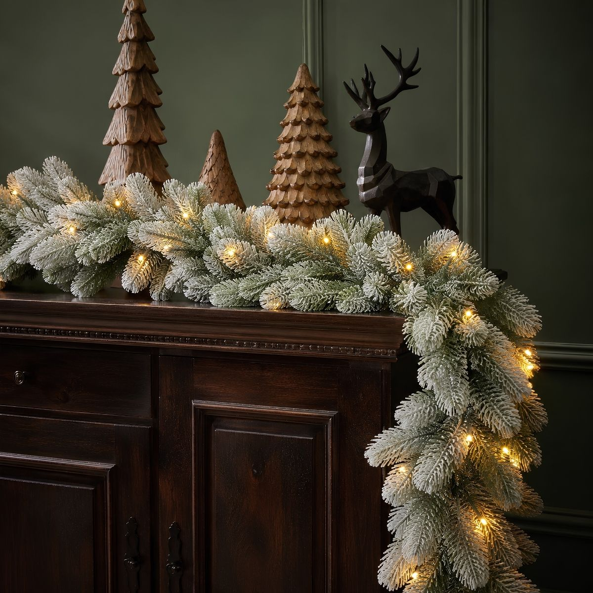 Verschneite grüne Weihnachtsgirlande mit warmem Licht auf dunklem Holzschrank mit Holz-Bäumen und Rentier.