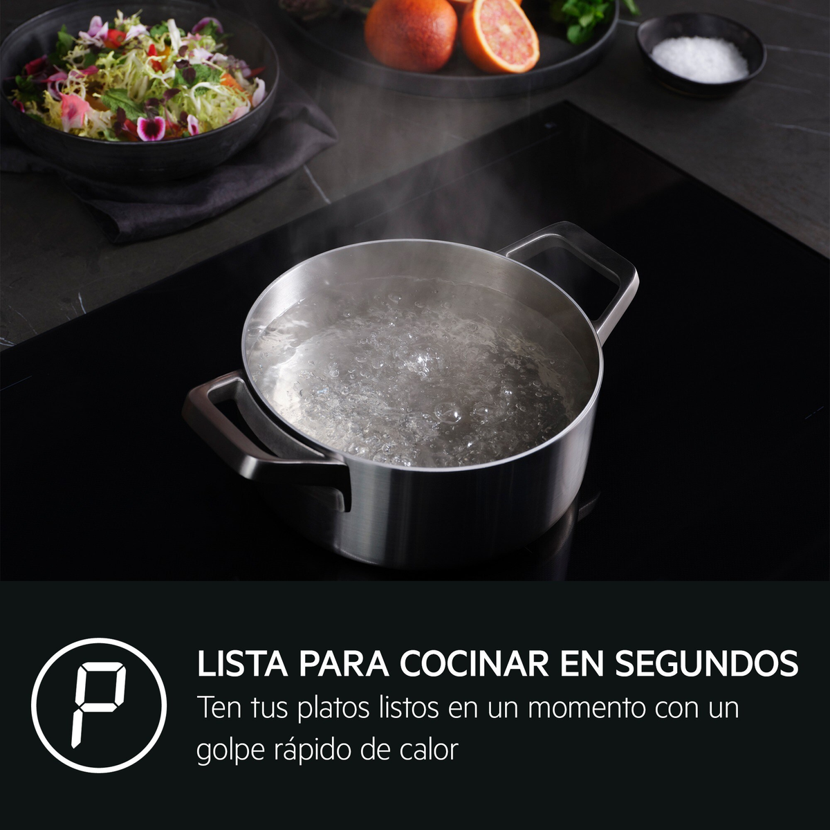 Olla de acero inoxidable con agua hirviendo en placa de inducción, con ensalada y cítricos al fondo.