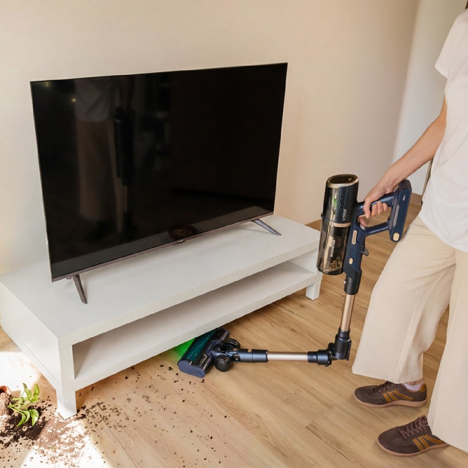 Una persona limpia la suciedad en un suelo de madera con una aspiradora de mano azul frente a un mueble de TV.