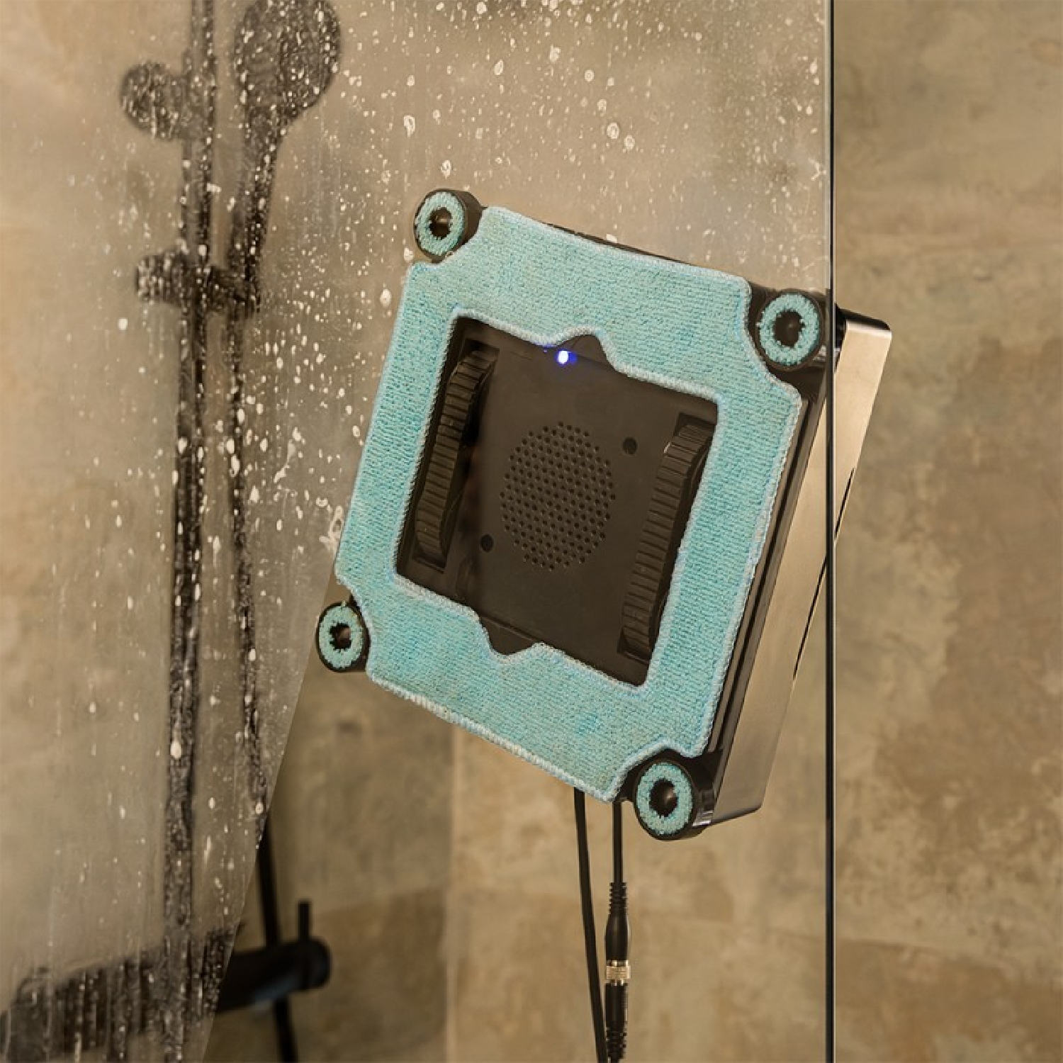 Un robot limpiacristales cuadrado con una almohadilla azul limpia una puerta de ducha con jabón.